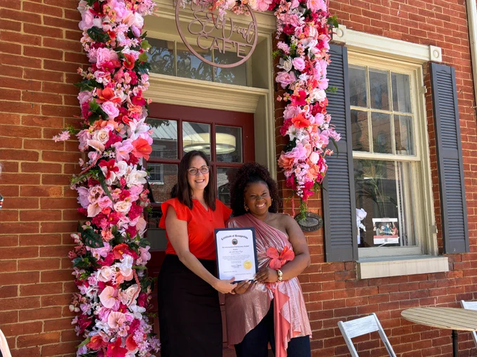 Two individuals stand in front of a brick building entrance decorated with a large floral display. The doorway is framed by an arch made of densely arranged artificial flowers in shades of pink, red, and white. Above the door hangs a circular sign featuring stylized text. One individual is wearing a bright red top and a dark skirt, while the other is dressed in a light, shimmery top with a draped design. Both individuals are holding a framed certificate together. To the right of the doorway is a tall window with blue shutters, and a small table with two metal chairs sits on the sidewalk in front of the building.