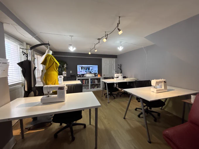 A well-organized sewing studio features several white worktables arranged around the room, each equipped with a sewing machine and a black rolling chair. The space has gray walls, wood flooring, and multiple ceiling-mounted track lights that illuminate the room evenly. On the left side, a dress form displays a bright yellow garment, positioned near a window with white blinds. Next to the dress form, a hanging rack holds fabric pieces. In the back of the room, a large TV screen shows an image of a sewing or craft workspace, and beneath it sits a shelving unit filled with books, supplies, and storage bins. Additional tables with sewing machines are arranged along the right side of the room. A red chair is partially visible in the lower right corner.