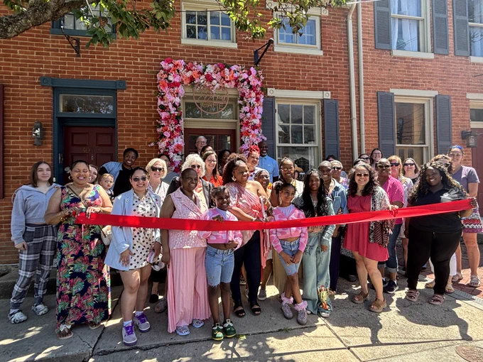 A large group of people stands closely together on a sidewalk in front of a brick building for a ribbon‑cutting ceremony. They are outdoors on a sunny day, gathered behind a long, bright red ribbon that stretches across the width of the group. The building behind them has red brick walls, white window trim, blue shutters, and a maroon door. At the center of the building’s façade, a doorway is decorated with an arch of pink, white, and red artificial flowers surrounding a sign above the entrance. Tree branches with green leaves extend across the upper left corner of the image, casting light shadows. The group is diverse in age and attire, wearing a mix of casual clothing, bright colors, patterns, and accessories.