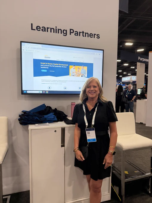 Missy Torence smiles at a trade show booth. A large screen displays \"Skyline: Invest in Human Capital Development and unlock the full potential of your workforce.\" Above the screen, a sign reads \"Learning Partners.\" Blue bags are on the counter.