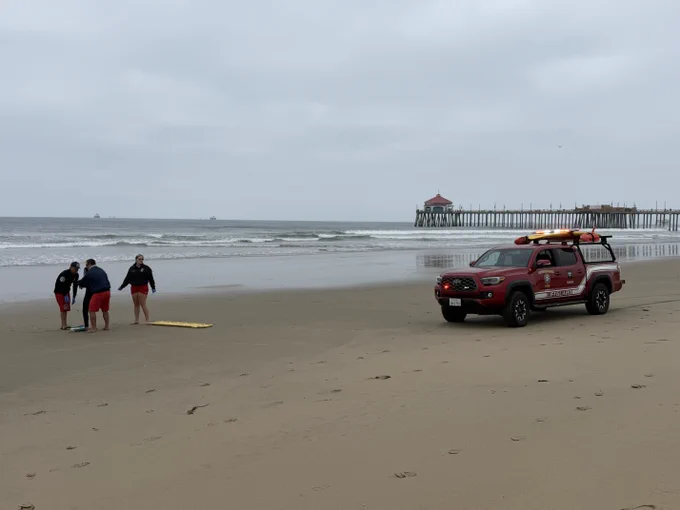 A Huntington Beach Fire Department Marine Safety rescue vehicle is parked on the sand near the shoreline while personnel prepare for a training exercise. The Huntington Beach Pier is visible in the background under overcast skies as waves break along the beach.