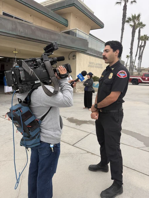 Dr. Rahimian, Chief Medical Officer for Huntington Beach Fire Department, speaks with a television news crew outside the Marine Safety Headquarters building. A cameraman records the interview as Dr. Rahimian discusses beach safety, pediatric trauma care, and the importance of emergency response training.