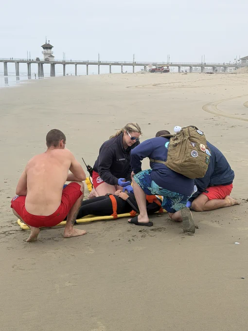 Huntington Beach Marine Safety personnel and medical responders provide care to a simulated pediatric trauma patient on the beach during a training exercise. Responders stabilize the patient on a backboard while coordinating treatment near the shoreline.