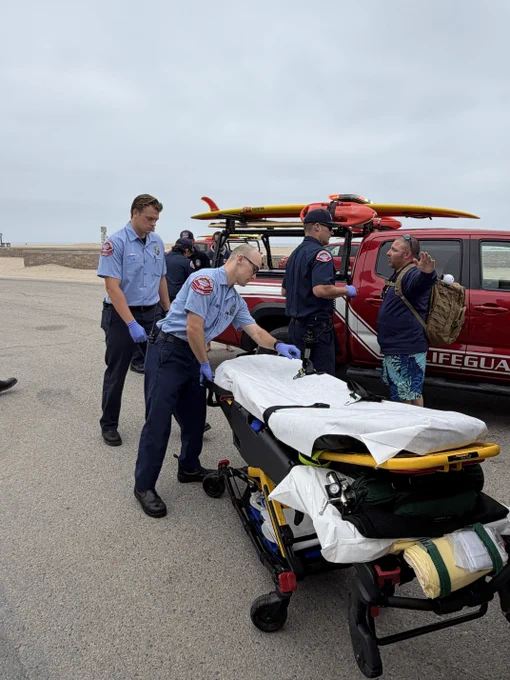 Firefighter paramedics and Marine Safety personnel prepare a gurney during a pediatric trauma training exercise near a Marine Safety rescue vehicle. Responders coordinate patient transport as part of a simulated beach emergency response.
