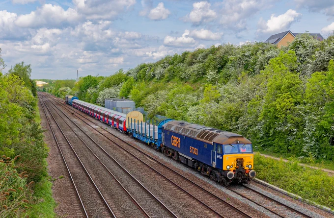 A freight train travels along a railway track surrounded by greenery and trees under a partly cloudy sky. Several other railway tracks run parallel to it.