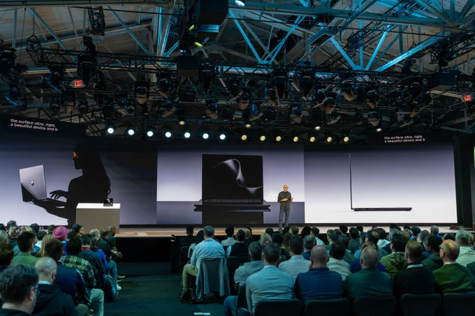 A speaker stands on stage in front of an audience, presenting a new device. The backdrop displays silhouette images of the laptop from different angles.