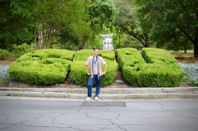 A person stands smiling in front of neatly trimmed hedges shaped into the letters "GSU," with lush trees and a pathway in the background.