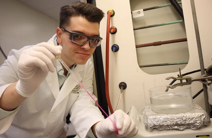 A person wearing safety glasses and a lab coat is handling a pipette in a laboratory setting. There are laboratory equipment and a container of liquid with aluminum foil nearby.