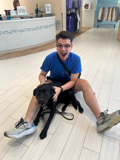 A person wearing glasses and a blue shirt is sitting on the floor. They are joyfully holding a black Labrador Retriever. The room has white tiled floors, clothes hanging in the background, and a counter with an "open" sign.