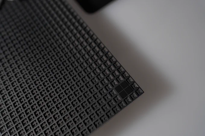 Close-up of a textured black grid surface with a square pattern, creating a three-dimensional appearance. The grid outlines are clearly defined, adding depth to the image.