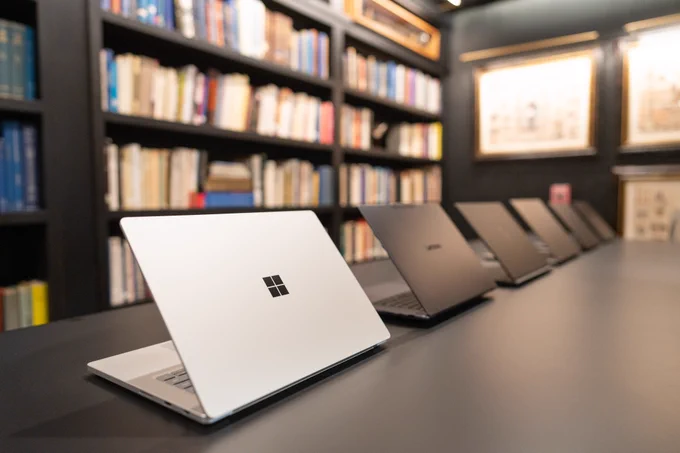 A row of devices on a dark table in a library setting, with a wall of bookshelves filled with books in the background. The closest device is a Microsoft Surface while the others are various dark colors.