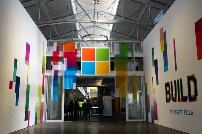 A spacious hall with a modern design featuring colorful banners in blue, green, red, and yellow. The Microsoft logo is prominently displayed at the top, and the word 'BUILD' is creatively styled on a wall.