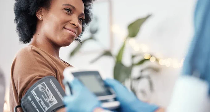 A person is having their blood pressure taken by a healthcare professional. They are smiling and wearing a brown shirt, while the professional wears blue gloves. The setting appears to be a well-lit, comfortable room with plants in the background.