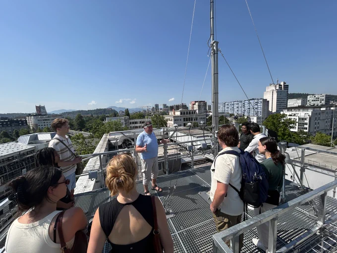 Water-R20 NRT students receive instruction on rooftop meteorological instrumentation at a Ljubljana weather monitoring facility, with the city skyline visible in the background.