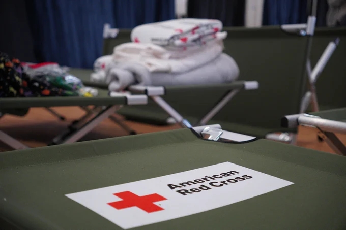 An array of American Red Cross cots with neatly folded blankets and pillows are arranged in a room, suggesting a disaster relief setup.