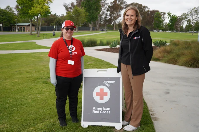 Two individuals stand outdoors near a sign with the American Red Cross logo. One person is wearing a red cap and shirt with the logo, and the other is in a black jacket. They are on a grassy area with trees and a building in the background.