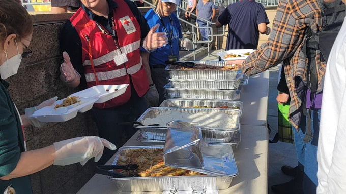 A group of people serving food from trays on a table, with some wearing Red Cross vests. A person in the foreground holds a foam container receiving food. 