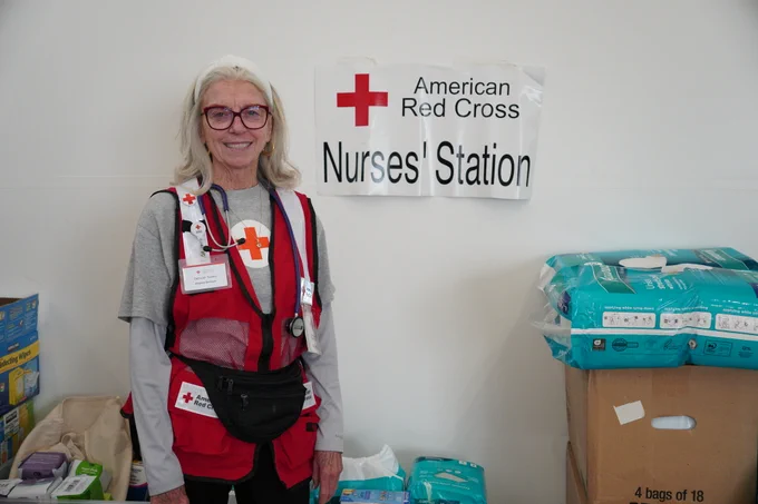 An individual wearing an American Red Cross vest stands in front of a "Nurses' Station" sign. The area is stocked with medical supplies, including boxes of diapers and other essentials.