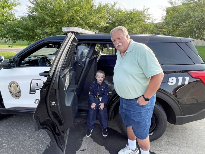 child wearing police costume sits inside back door of a Police cruiser and adult stands next to them