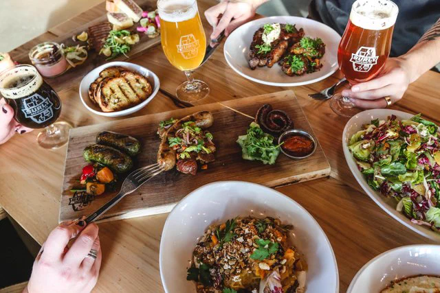 A table at Surly Brewing Co filled with various dishes, including grilled meats, a fresh salad, and a bowl of pasta. There are three glasses of beer, each with a unique color. A few hands are reaching for food and drinks.