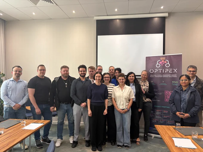 Image shows the OptiPEx consortium standing by the project banner in front of a projector.