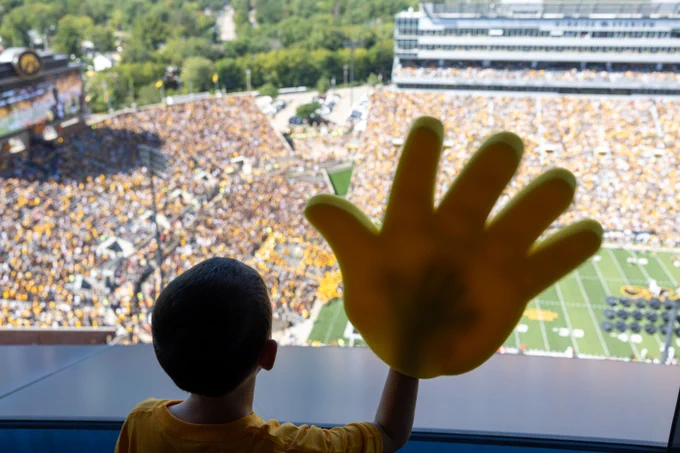 A child, wearing a yellow shirt, stands inside a stadium suite, looking out over a packed football stadium. The child raises a large yellow foam hand in front of a window, with the green football field and colorful crowd visible in the background.