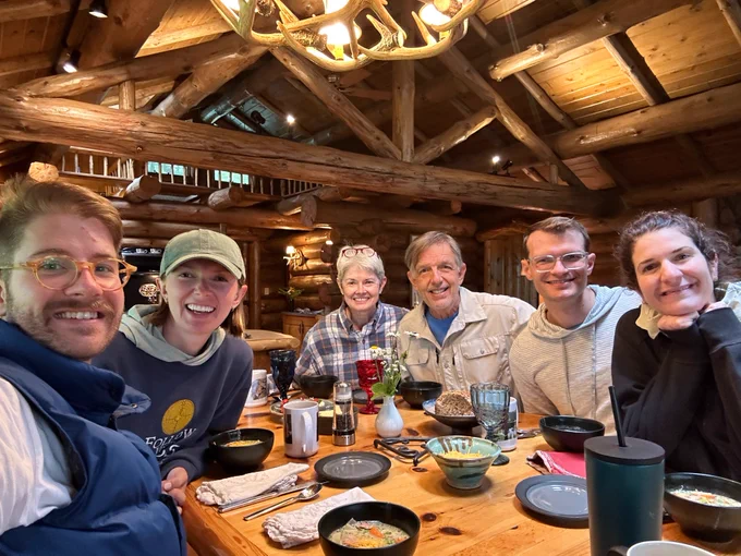 A group of six people sits around a wooden table inside a rustic log cabin. The table is set with bowls of food and drinks. Above them, an antler chandelier hangs from the ceiling. The atmosphere is cozy and warm, with everyone smiling at the camera.