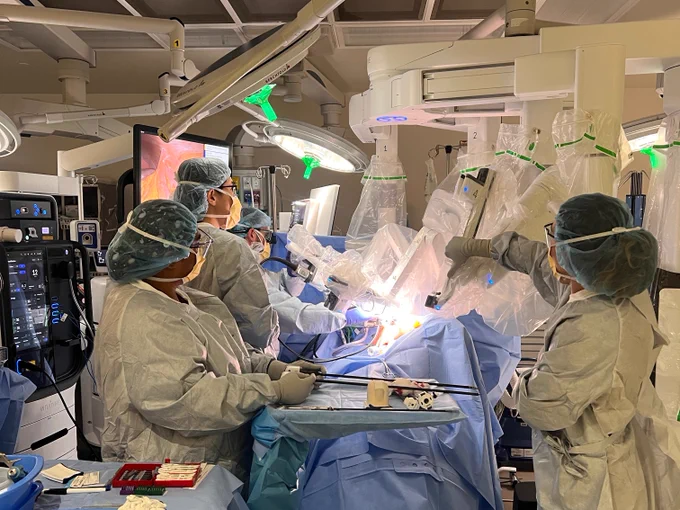 A team of surgeons in a modern operating room performs surgery with robotic assistance. The medical staff, wearing blue scrubs, gloves, and masks, surrounds the patient, who is covered with blue sterile drapes. Robotic arms, covered in plastic, assist in the procedure. Bright surgical lights illuminate the area, and monitors display surgical visuals. Surgical instruments and equipment are organized on nearby tables.