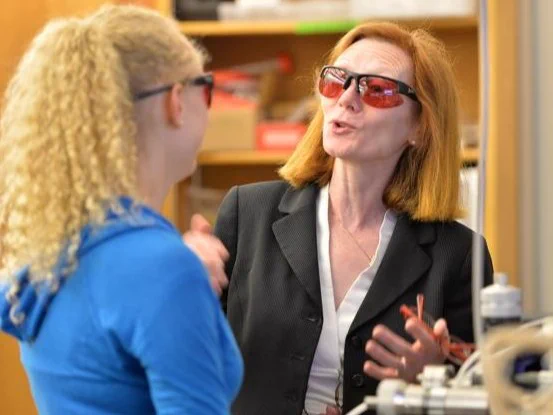 Margaret Murnane and another individual wearing safety goggles engaged in conversation in a lab setting with scientific equipment visible in the foreground.