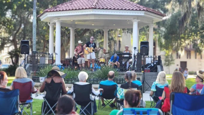 A small band performs on a gazebo stage in a park. The audience sits on foldable chairs and blankets on the grass. The atmosphere is relaxed, with trees and historic buildings in the background.