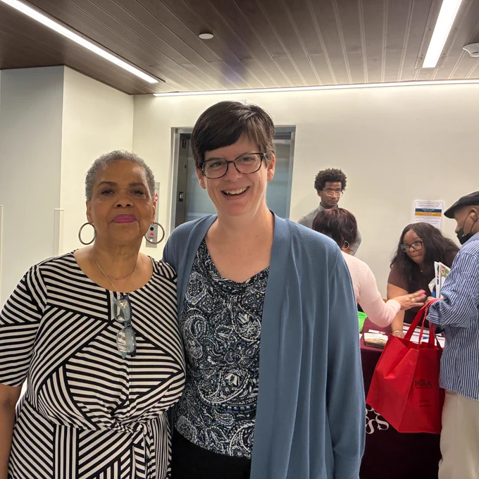 Angela Telesford (left), program coordinator at NCBA, and Kari Benson, Deputy Assistant Secretary for Aging, pose smiling in front of an information table where people are picking up materials and conversing