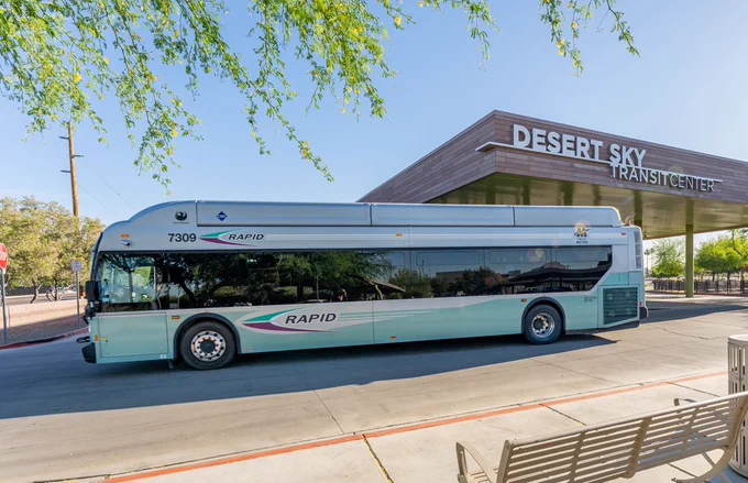 A Phoenix RAPID bus is parked in front of Desert Sky Transit Center. The scene is sunny, with trees providing shade. A bench is visible in the foreground.