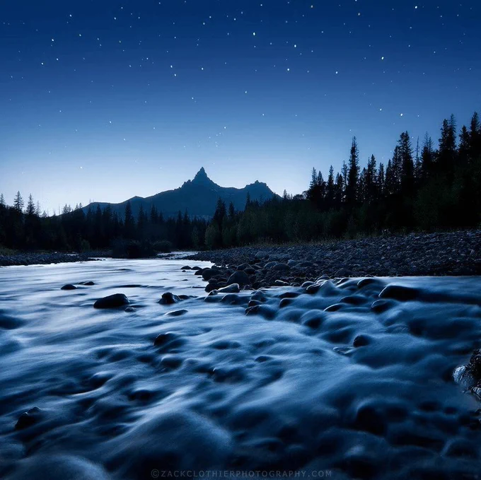 A river flows over rocks with pine trees and mountains in the background, set beneath a clear night sky filled with stars, and the deep indigo shades of blue hour.