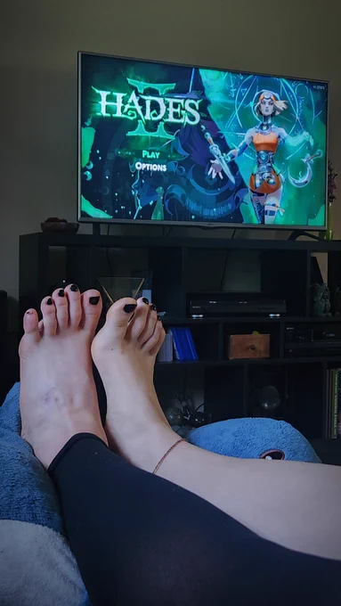my feet resting on a blahaj stuffed shark in front of a TV with the Hades 2 main menu on it.