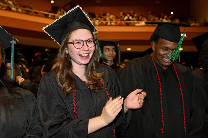 A group of graduates in black caps and gowns, adorned with red cords, celebrate at a graduation ceremony. One individual at the front is clapping joyfully, surrounded by peers. In the background, an auditorium filled with audience members can be seen.