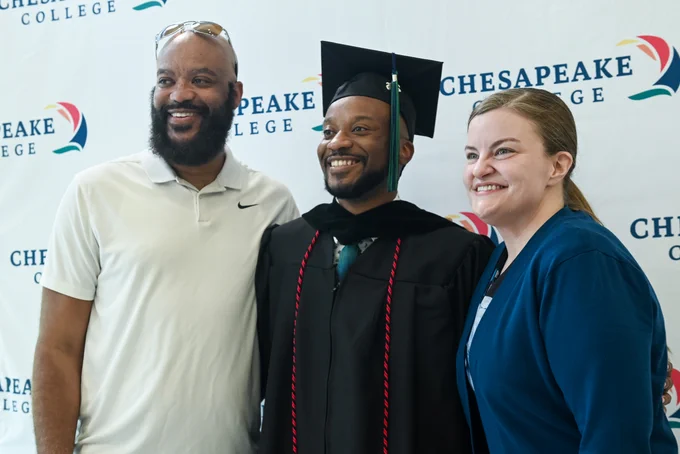 A graduate wearing a black cap and gown smiles while standing between two people. They are in front of a backdrop with the Chesapeake College logo repeated on it.