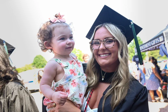 A person in graduation attire and cap is smiling and holding a young child with curly hair and floral dress. The scene appears to be outdoors with people and a tent in the background.
