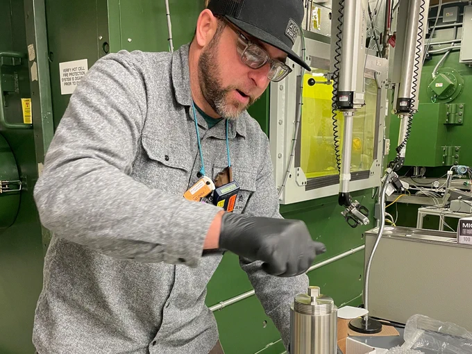 Jamin Trevino practicing the steps for handling the special form capsule that holds the radium-226. He has a metal cylinder in his left hand and various equipment for handling radioactive material behind him. 
