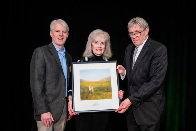 Three professionally dressed people posing for a picture. The woman in the middle is holding a framed picture of a painted deer.