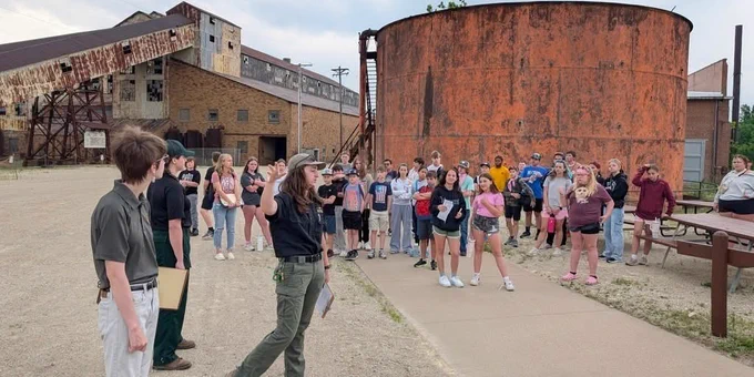 Group of young people gathered outdoors near large rusted industrial tanks and buildings on a cloudy day.