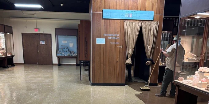 Man sweeping floor near a wooden booth with curtains inside a museum gallery room.