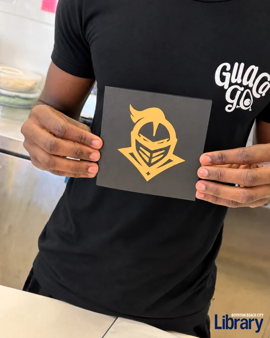 Person holding a black card featuring a gold knight helmet logo, wearing a black shirt with white text, standing indoors near a counter.