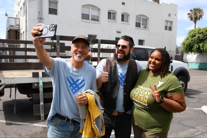 Three smiling adults, one holding a phone, pose for a selfie outdoors in front of a building and a parked truck.
