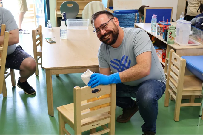 Smiling man wearing blue gloves cleans a small wooden chair in a classroom with tables and children's water bottles.