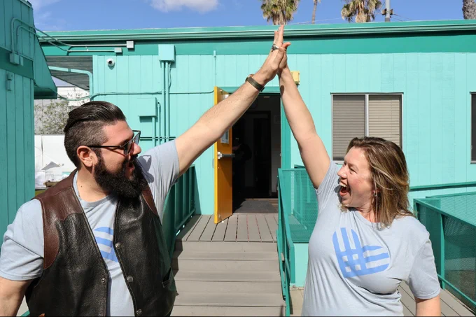 Two smiling adults high-five enthusiastically on a wooden walkway outside a turquoise building