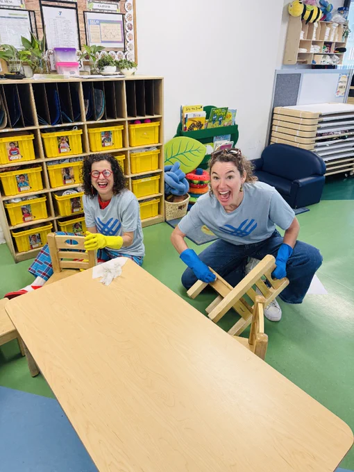 Two smiling adults in gloves cleaning and stacking small wooden chairs in a colorful preschool classroom.