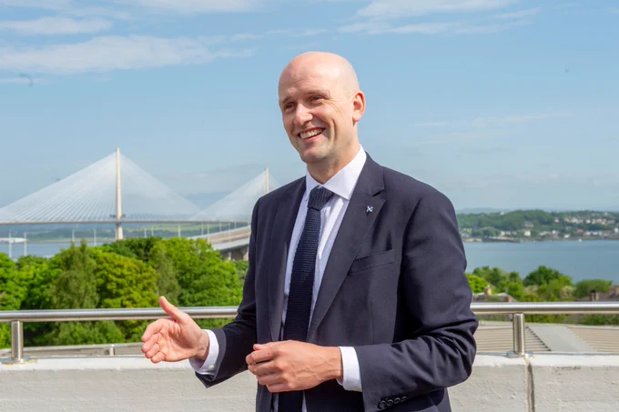 Transport Secretary smiles on camera, on a sunny day with the South Queensferry Crossing in the background.