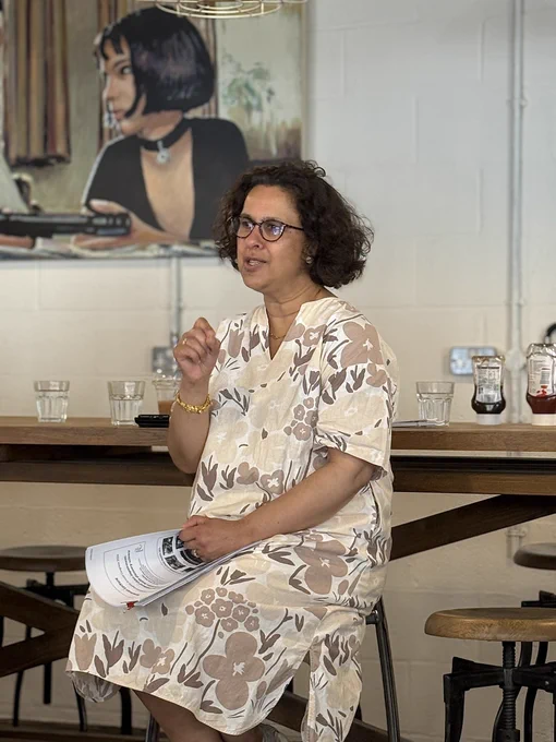 Prof Jugnoo Rahi is seated on a high stool speaking at a coffee hour event, holding printed notes. She is in a café with wooden tables, water glasses, and condiment bottles. A large piece of artwork featuring a person is visible on the wall behind.