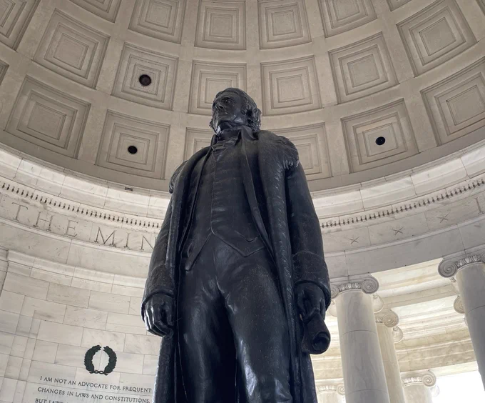 A tall bronze statue of Thomas Jefferson wearing a long coat stands in the center of a round memorial chamber.