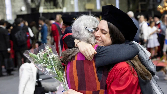 Two individuals embrace at the 2026 Cornell PhD Recognition Ceremony. One person wears a graduation cap and gown, holding a bouquet of flowers. The background shows others in similar academic attire.